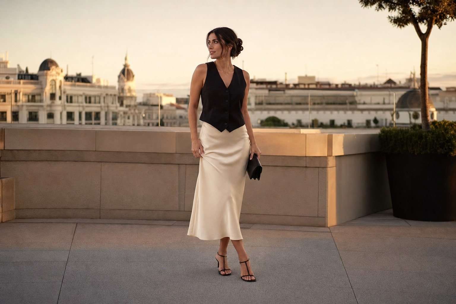 Mujer con chaleco negro y falda satinada marfil en una terraza al atardecer.